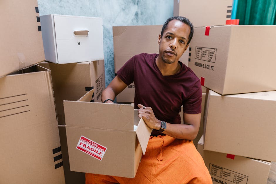 A man with short dark hair, wearing a maroon T-shirt, orange pants, and a smartwatch, is inside a room surrounded by multiple cardboard boxes used for home relocation. He is sitting on the floor, hold