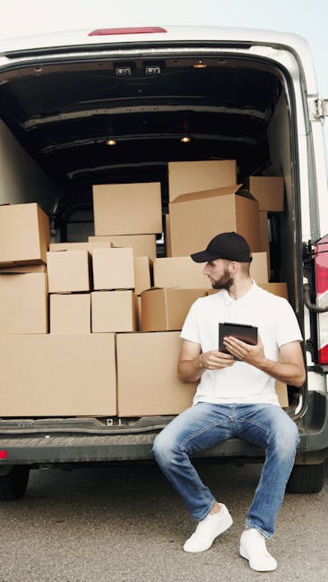 A man wearing a black cap, white t-shirt, and blue jeans is seated on the edge of an open van loaded with numerous cardboard boxes of various sizes, some sealed with packing tape and others open with visible packing paper or plastic wrap. The van is parked outside a property, and the boxes are arranged inside in preparation for a home relocation or furniture transport. The interior of the van shows a dark ceiling with visible hooks and attachments for securing cargo. The scene captures part of a loading process involving packing materials like cardboard boxes for house removals, with the man referencing a portable device, possibly coordinating the move or checking inventory. The background outside is neutral, emphasizing the focus on the loading activity and furniture or goods being transported by Bermondsey Man and Van, a company offering quick local removals in SE16 for house moves and relocation services.