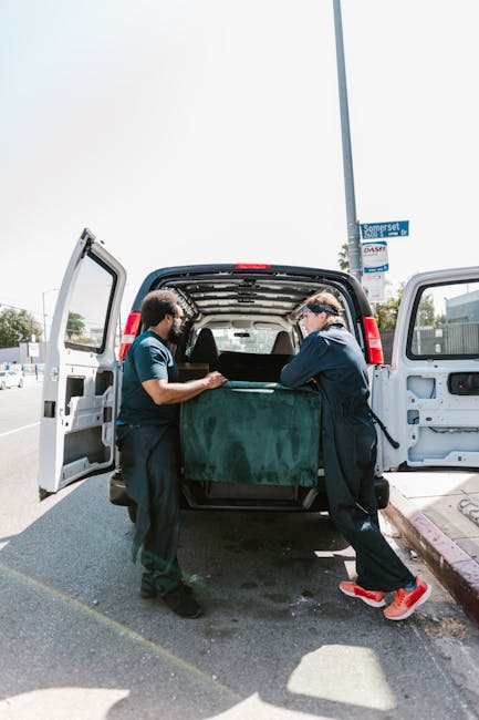 Two movers from Bermondsey Man and Van are engaged in a home relocation task, loading a large piece of furniture wrapped in dark green protective fabric into the back of a black van. The scene takes place on a paved street during daylight, with the open rear doors of the van revealing a spacious cargo area. One mover, wearing a dark blue uniform and a headband, is securing the furniture as the other, in a similar uniform with a teal shirt, assists with guiding and lifting the item. Inside the van, visible within the cargo space, are additional packing materials such as cardboard boxes and plastic wrap, indicating careful packing for transportation. A tall street sign and some buildings are visible in the background, situating the activity in an urban environment. This image illustrates professional furniture transport and packing during a local house removal, highlighting the logistical process managed by Bermondsey Man and Van as part of their removals service category.