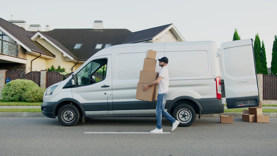 A man wearing a black cap, white t-shirt, and blue jeans is seated on the edge of an open van loaded with numerous cardboard boxes of various sizes, some sealed with packing tape and others open with visible packing paper or plastic wrap. The van is parked outside a property, and the boxes are arranged inside in preparation for a home relocation or furniture transport. The interior of the van shows a dark ceiling with visible hooks and attachments for securing cargo. The scene captures part of a loading process involving packing materials like cardboard boxes for house removals, with the man referencing a portable device, possibly coordinating the move or checking inventory. The background outside is neutral, emphasizing the focus on the loading activity and furniture or goods being transported by Bermondsey Man and Van, a company offering quick local removals in SE16 for house moves and relocation services.