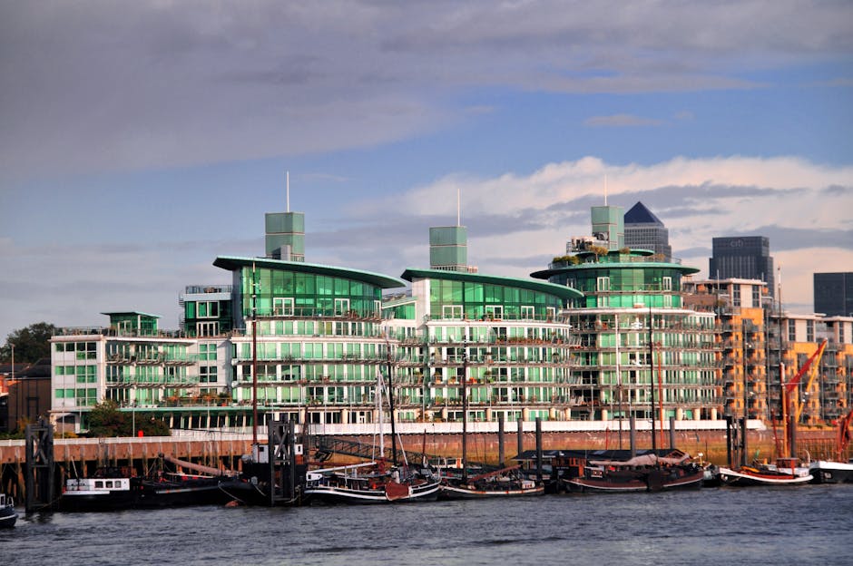 A modern, multi-storey building with curved green-tinted glass facades and flat, asymmetrical roofs, situated along a waterfront with a row of boats moored at the dock. The building features numerous 