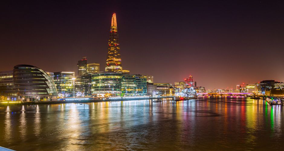 Nighttime view of London's skyline featuring the illuminated Shard skyscraper prominently rising above other modern office buildings with glass facades; reflective river in the foreground shows the colorful city lights, including shades of yellow, red, and green, shimmering on the water's surface; the scene is calm with clear visibility of the waterfront area, which may be part of a residential or commercial estate; lighting highlights the sleek architectural designs characteristic of central London; the image captures the vibrant urban environment typical of a home relocation or furniture transport process in the city, with no visible moving activity but emphasizing the cityscape associated with Bermondsey and Southwark areas in London.