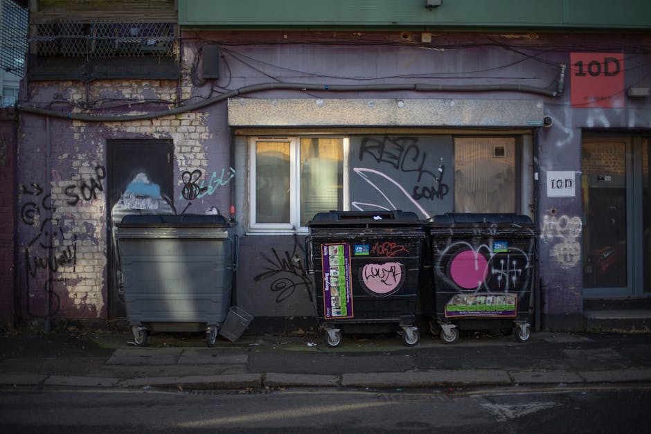 A row of three large wheeled refuse bins positioned on the pavement in front of a building with a brick and concrete exterior. The left bin is grey with a closed lid, while the two black bins next to it have graffiti and stickers on their surfaces, including a pink heart and various tags. The building behind features a small window with closed blinds and walls covered with colorful graffiti, including tags, illustrations, and spray-painted designs. A metal conduit runs horizontally above the bins, and a red sign with the number '10D' is visible. The scene appears to be an urban area with a mix of residential and commercial elements, depicting a typical home removal or packing environment with waste disposal features, indicative of ongoing moving or clearance activities, often coordinated by companies such as Bermondsey Man and Van.